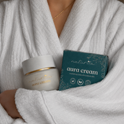 Person wrapped in a white robe with aura cream jar and box on a neutral background